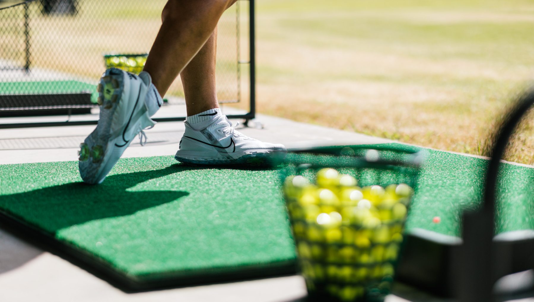 Close-up of a person in white sneakers stepping onto a green court beside a basket of tennis balls.
