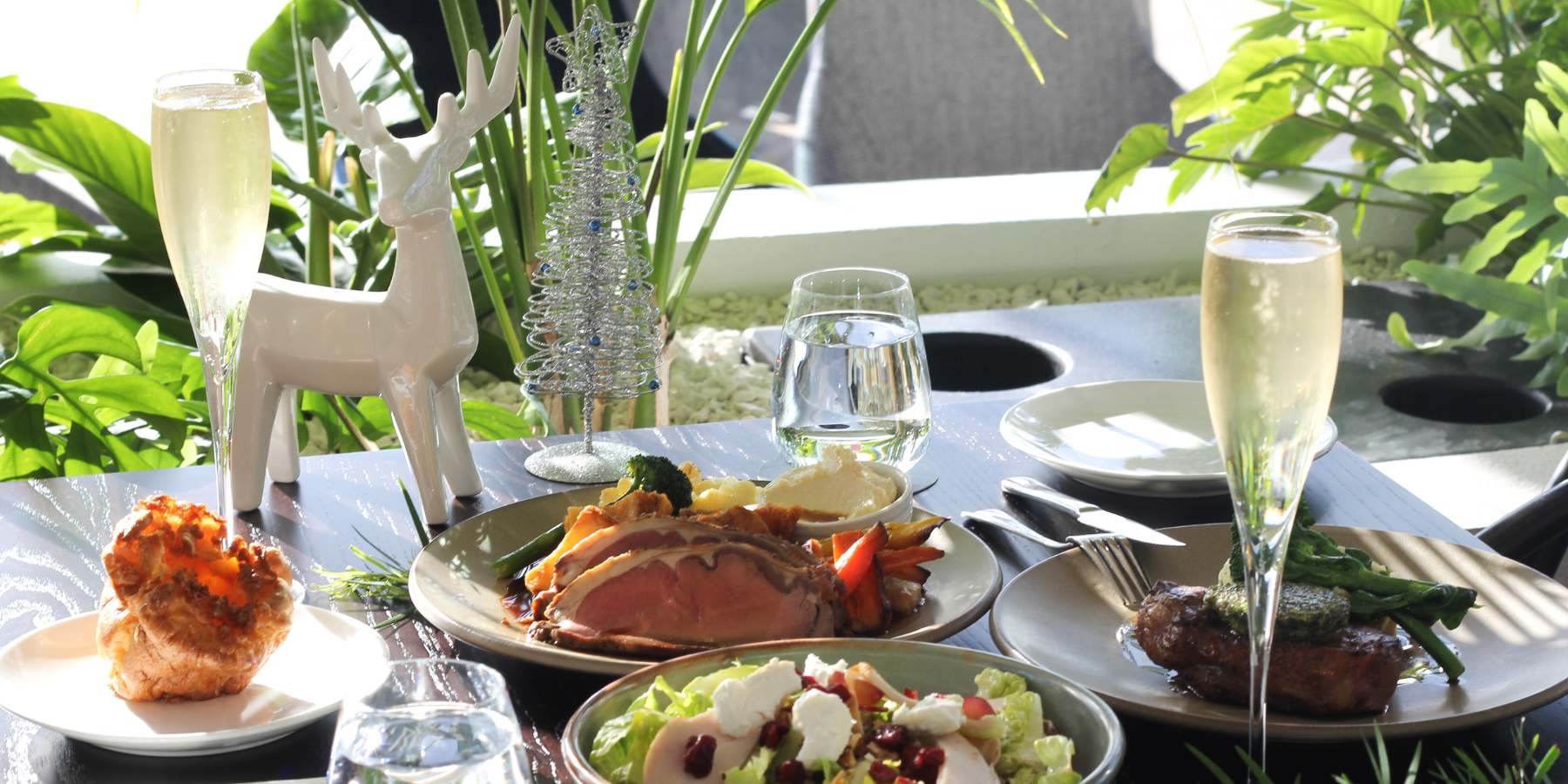 A sunlit dining table set with roast meats, salad, champagne flutes, water glasses, and white deer and tree decorations among leafy plants.