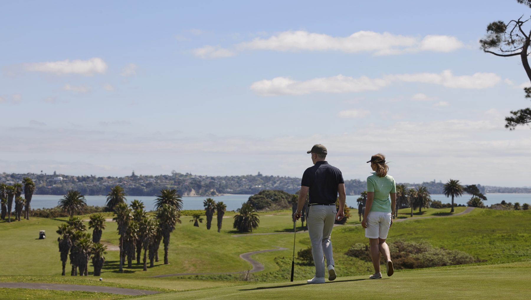 Two golfers walk across a green course lined with palm trees, overlooking the ocean and a coastal city.