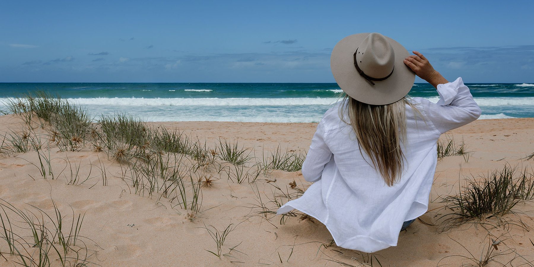 A woman in a white shirt and hat sits on a sandy beach, gazing at the waves and ocean under a blue sky.