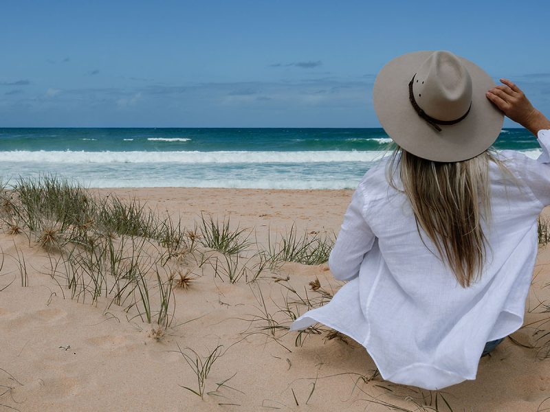 A woman in a white shirt and hat sits on a sandy beach, gazing at the waves and ocean under a blue sky.