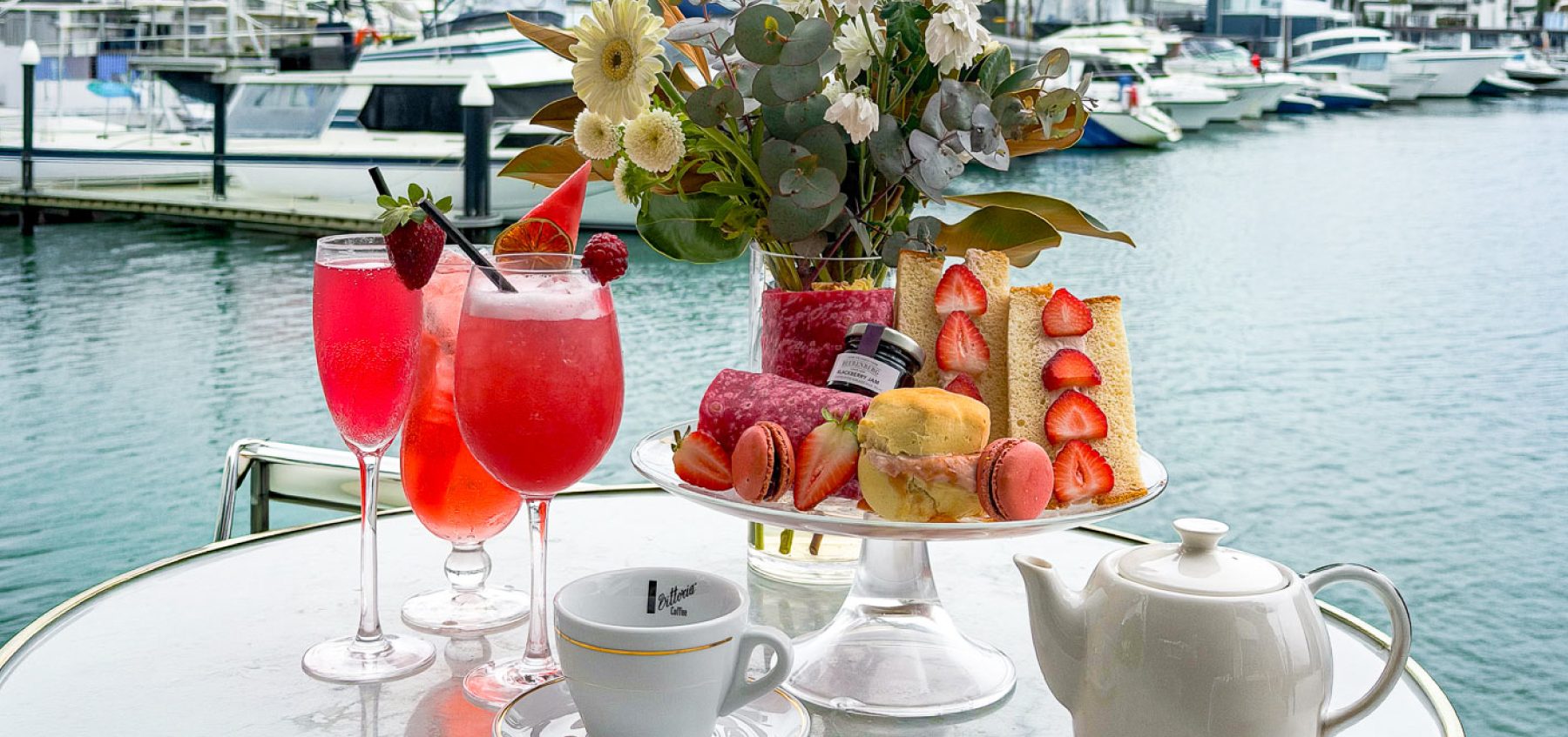 Brightly colored cocktails and a variety of pastries are arranged on a table overlooking a marina with boats. A teapot and a decorative floral arrangement complete the setting.