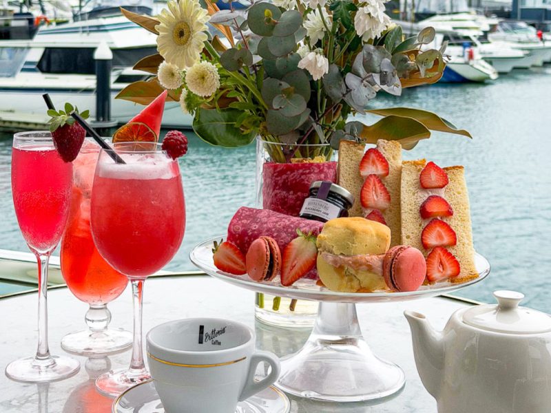 Brightly colored cocktails and a variety of pastries are arranged on a table overlooking a marina with boats. A teapot and a decorative floral arrangement complete the setting.