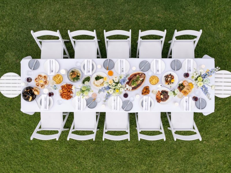 Overhead view of a long outdoor dining table set with plates, assorted food, flowers, and white chairs on a lawn.