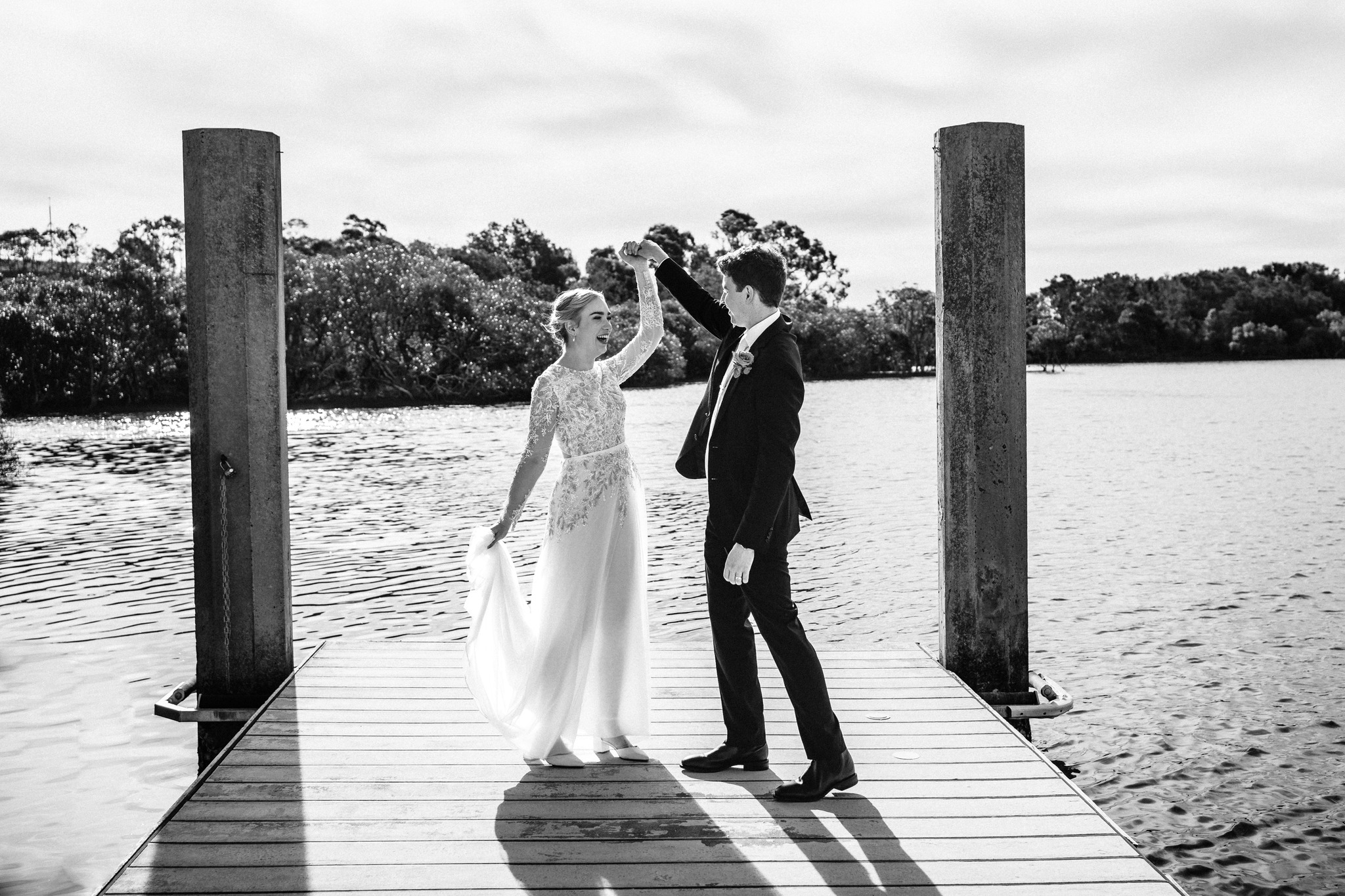 A bride and groom smile as they dance on a wooden dock over the water, framed by tall posts.