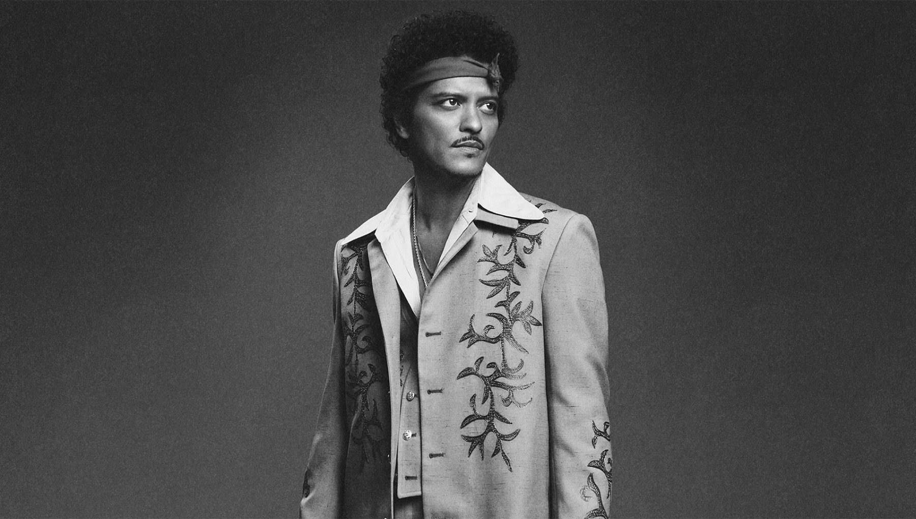 Black-and-white portrait of a curly-haired man wearing a headband and an embroidered suit, gazing off to the side.