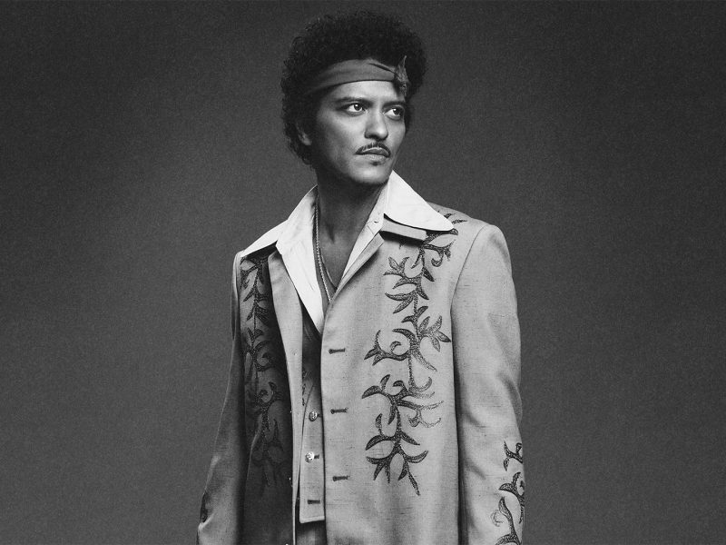 Black-and-white portrait of a curly-haired man wearing a headband and an embroidered suit, gazing off to the side.