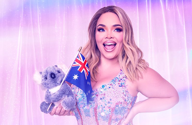 Smiling woman in a sparkling dress holds a koala plush toy and Australian flag against a pink curtain.