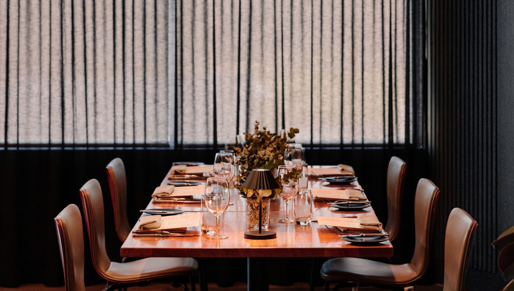 Private dining table set for eight at Gather North in Hunter Valley, featuring elegant place settings, wine glasses, floral centerpiece, and ambient lighting, framed by tall windows with vertical blinds.