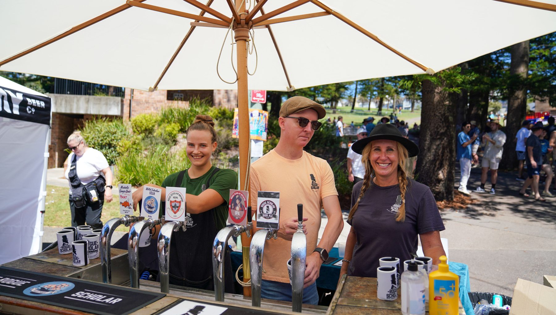 Three people stand behind a beer stall with taps and cups beneath a large white umbrella at an outdoor festival.