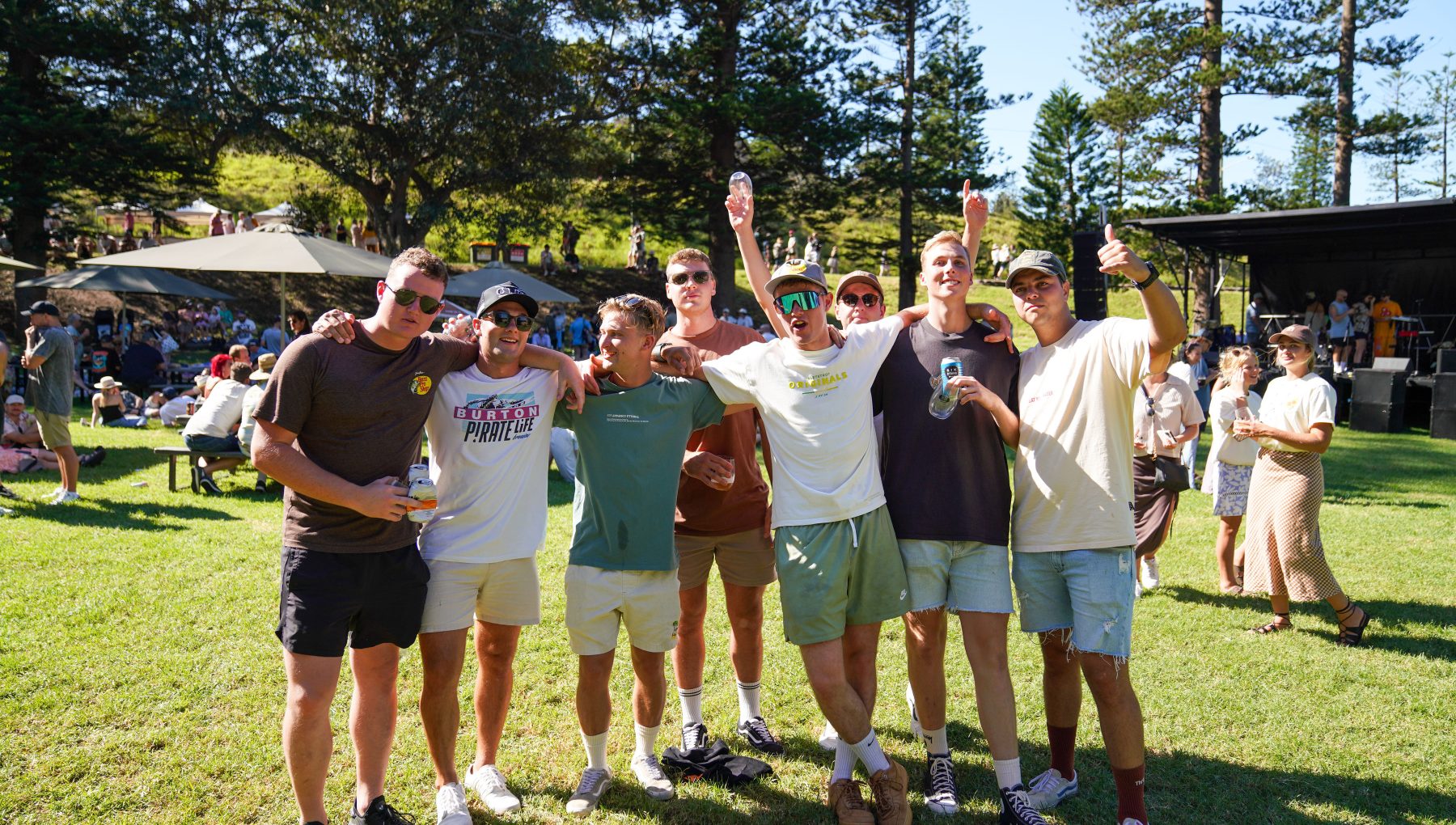 A group of young men pose with arms around each other at a sunny outdoor gathering, raising hands and holding drinks.