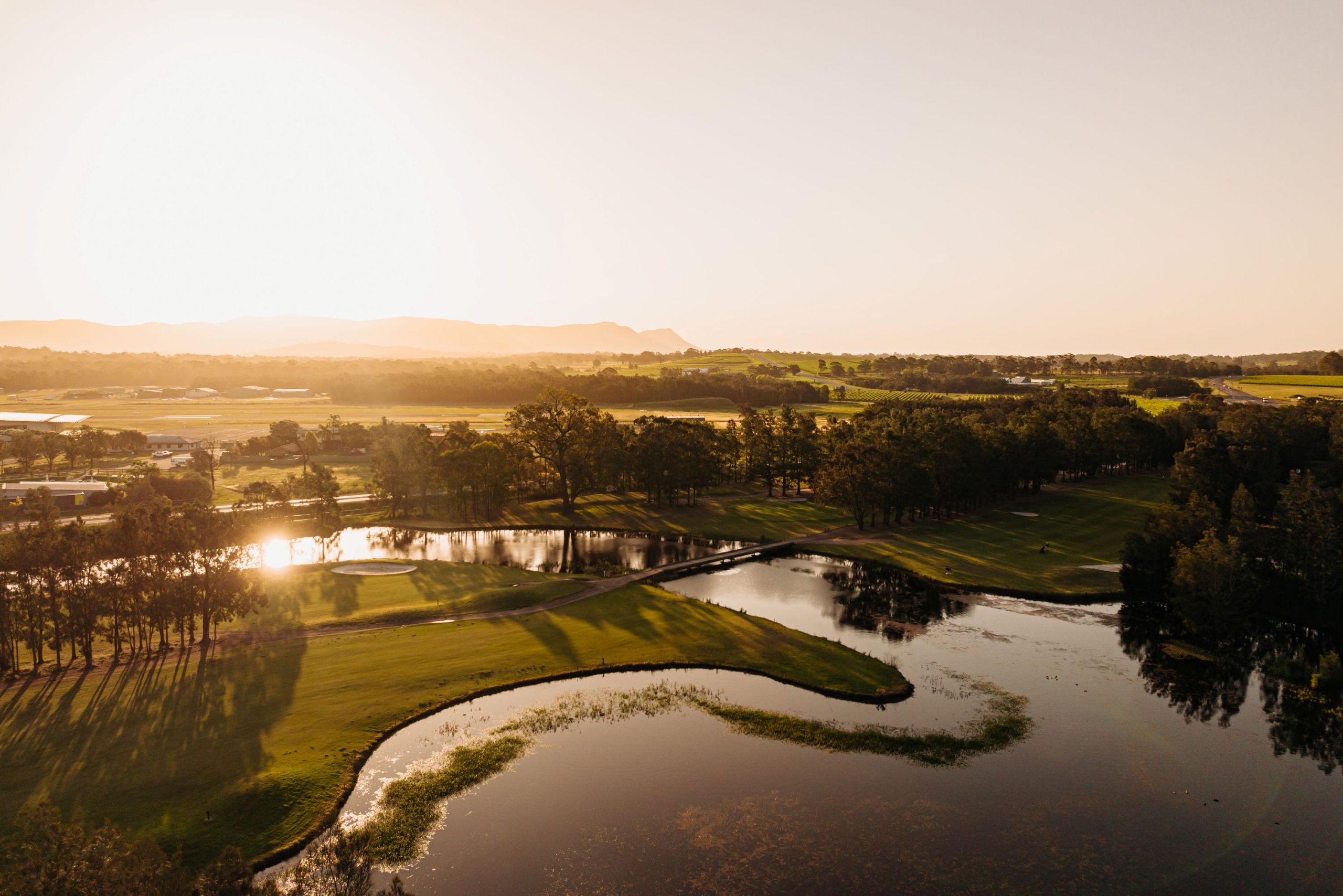 Golden-hour aerial view of a golf course with ponds and long tree shadows, with fields and hazy hills in the distance.
