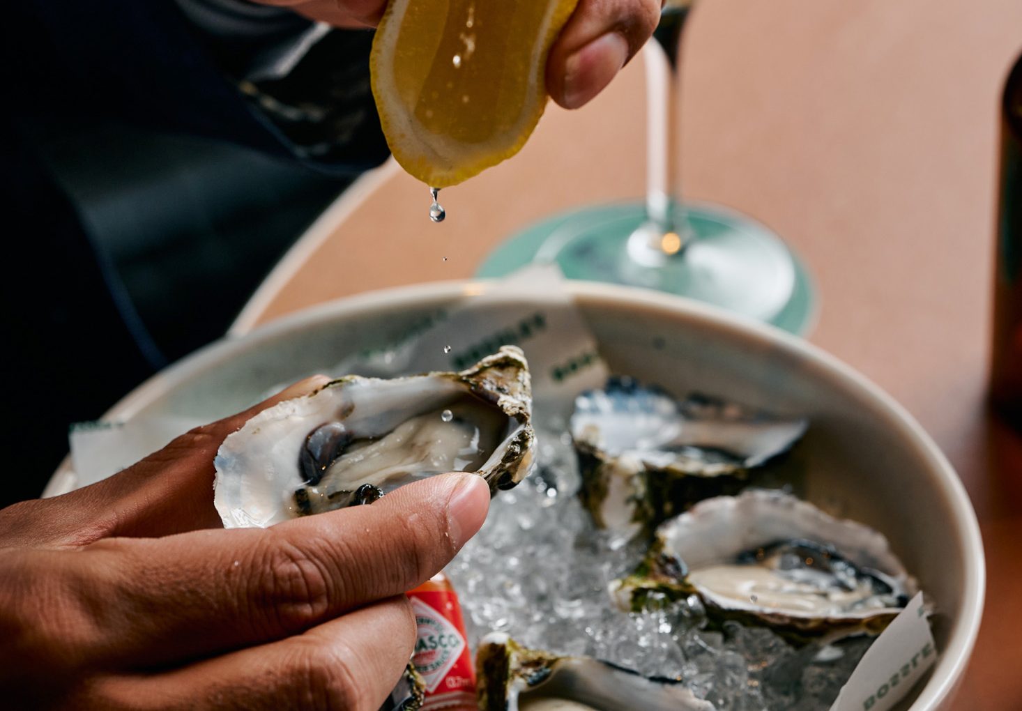 A hand squeezes a lemon over a plate of oysters on ice, with a bottle of hot sauce visible nearby.