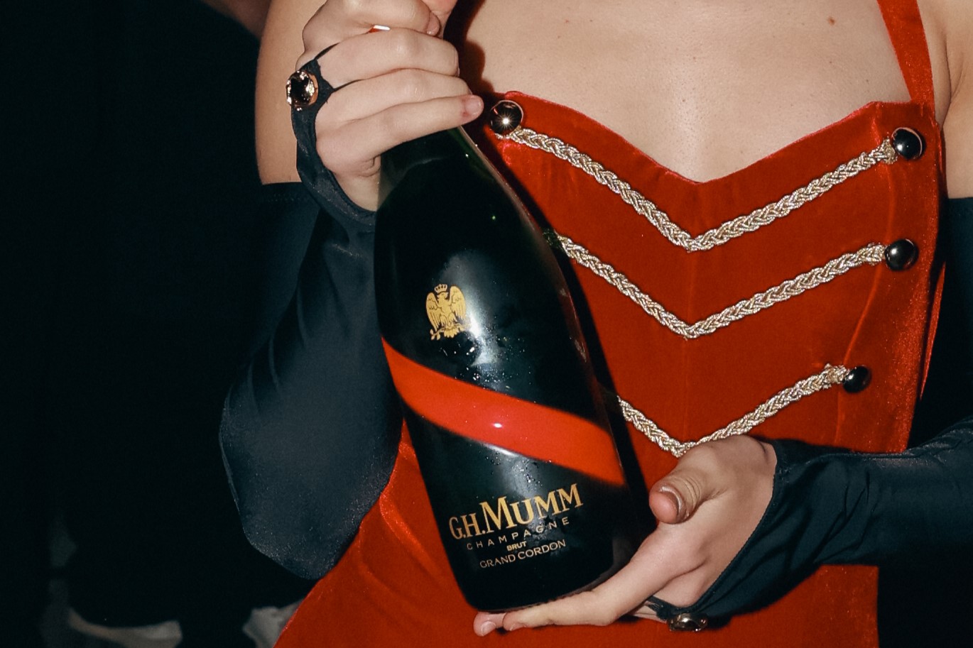 A person in a red jacket with silver trim holds a large black champagne bottle with a red stripe.