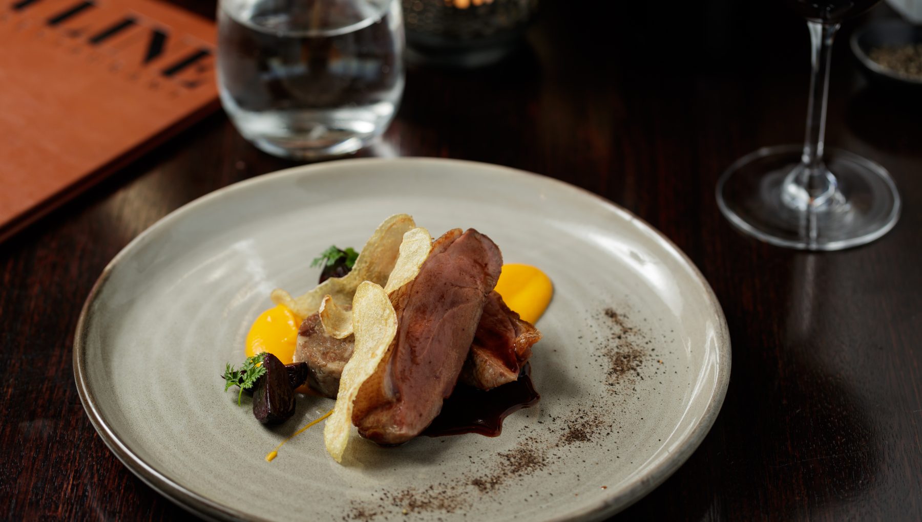 Sliced roast meat topped with crisp potato slices and herbs, with yellow sauce on a ceramic plate.
