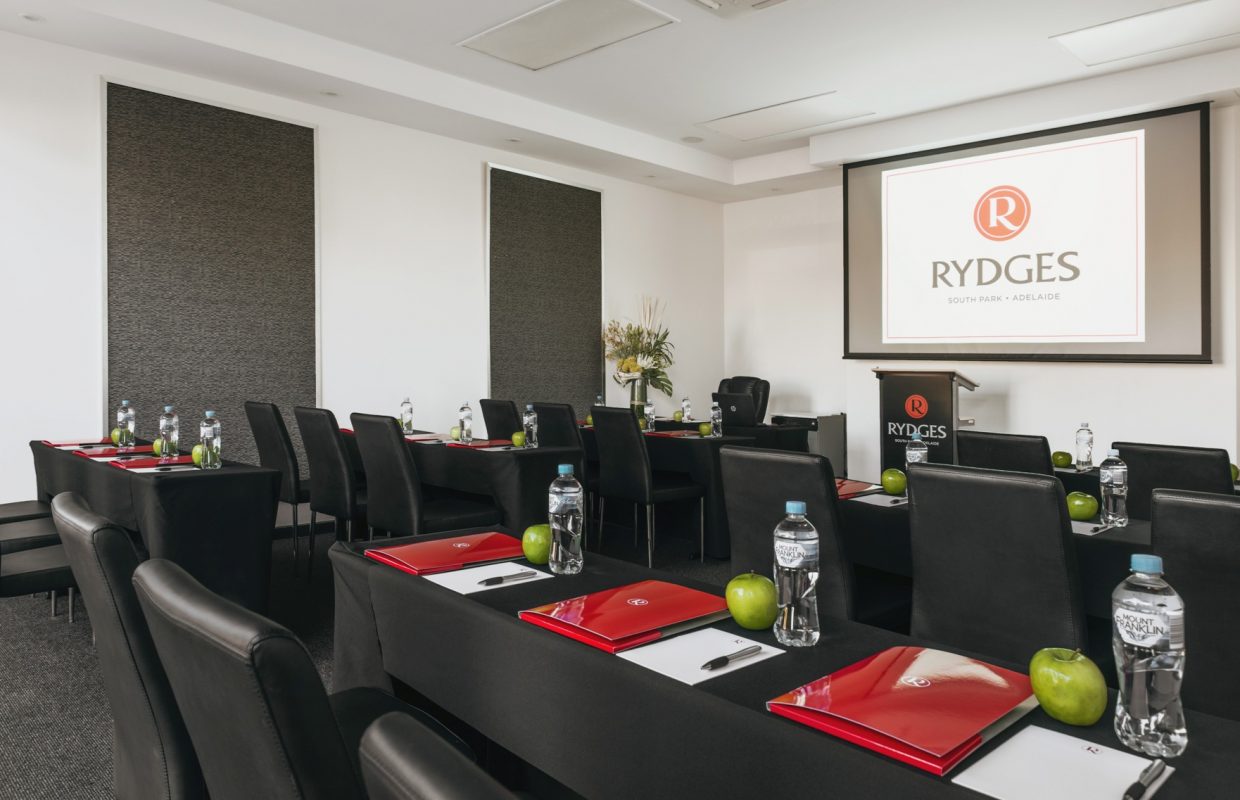 A modern conference room with black tables and chairs, featuring red folders, water bottles, and green apples at each setting. A projector screen displays the Rydges logo.