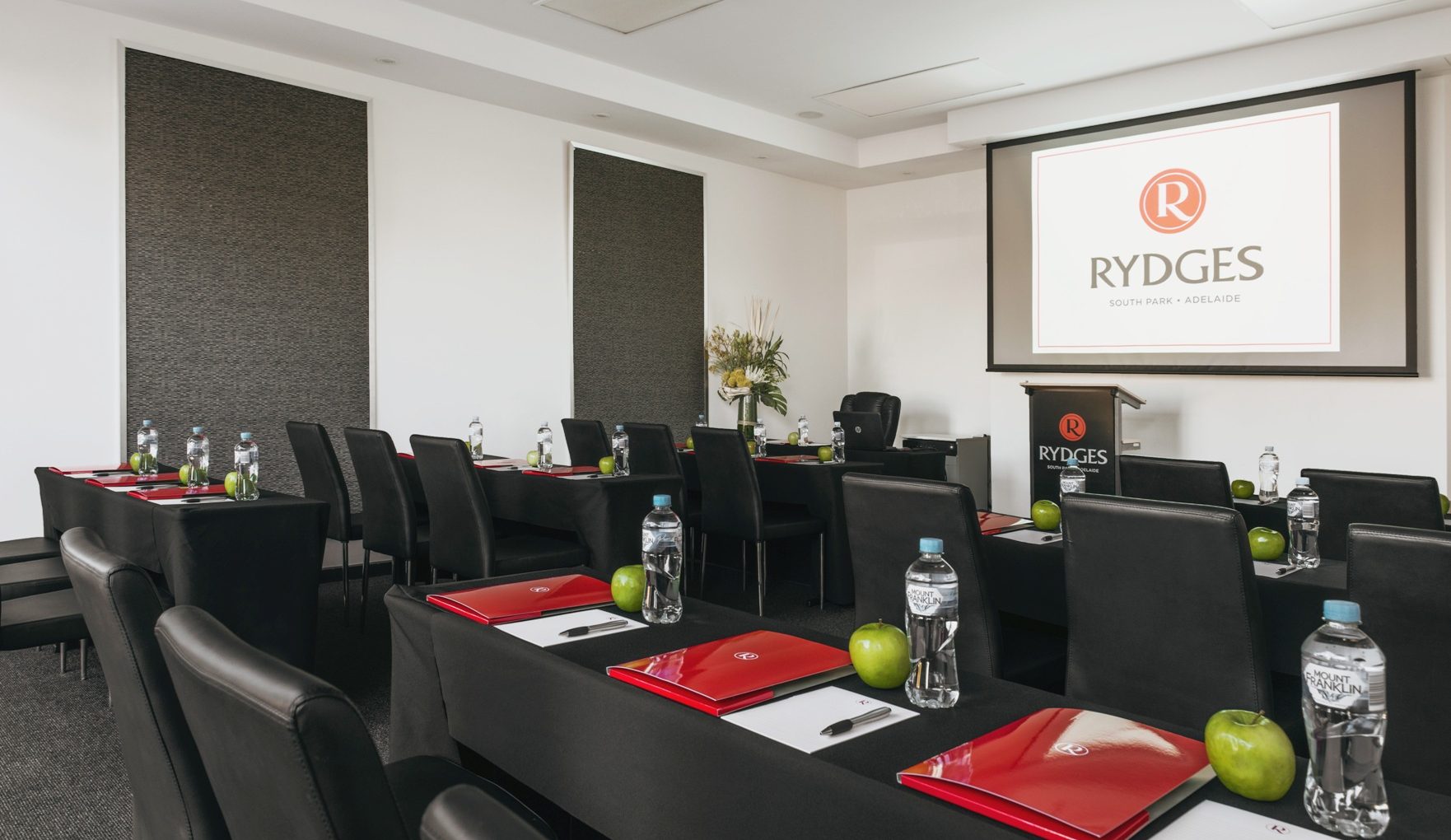 A modern conference room with black tables and chairs, featuring red folders, water bottles, and green apples at each setting. A projector screen displays the Rydges logo.