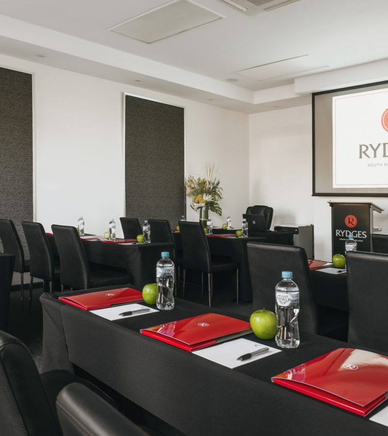 A modern conference room with black tables and chairs, featuring red folders, water bottles, and green apples at each setting. A projector screen displays the Rydges logo.