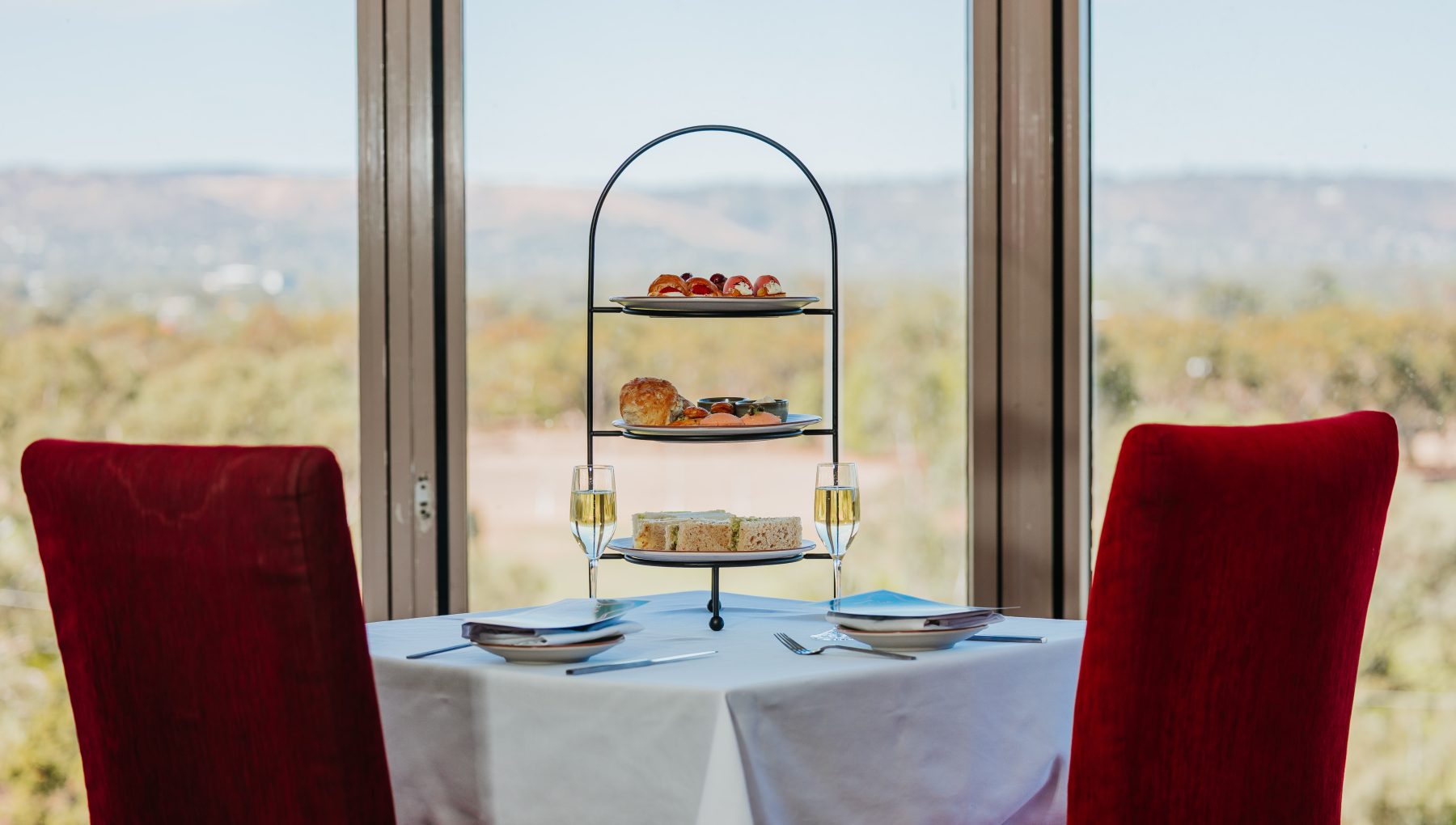 A neatly set table with red chairs features an elegant three-tier stand filled with assorted pastries and two glasses of champagne, framed by large windows showcasing the outdoors.