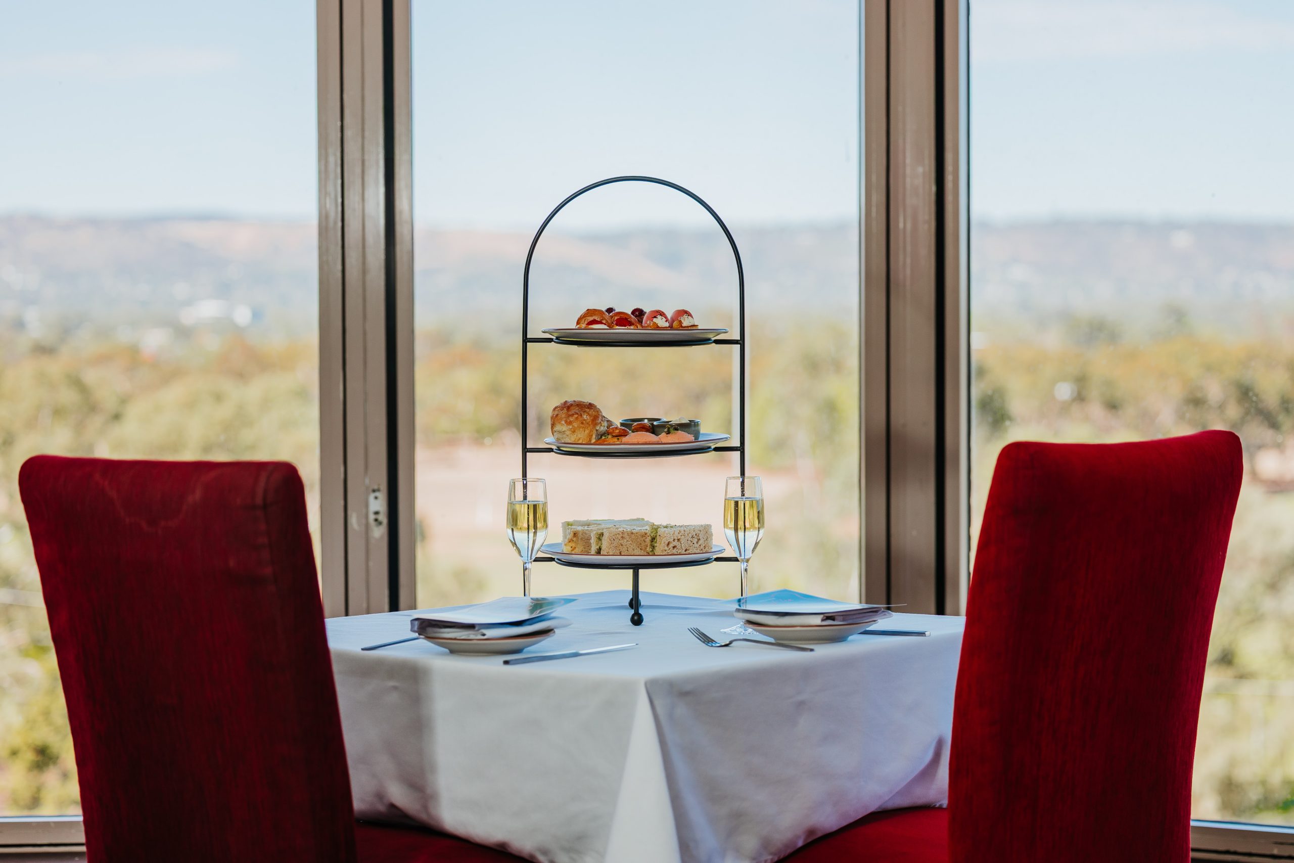 A neatly set table with red chairs features an elegant three-tier stand filled with assorted pastries and two glasses of champagne, framed by large windows showcasing the outdoors.