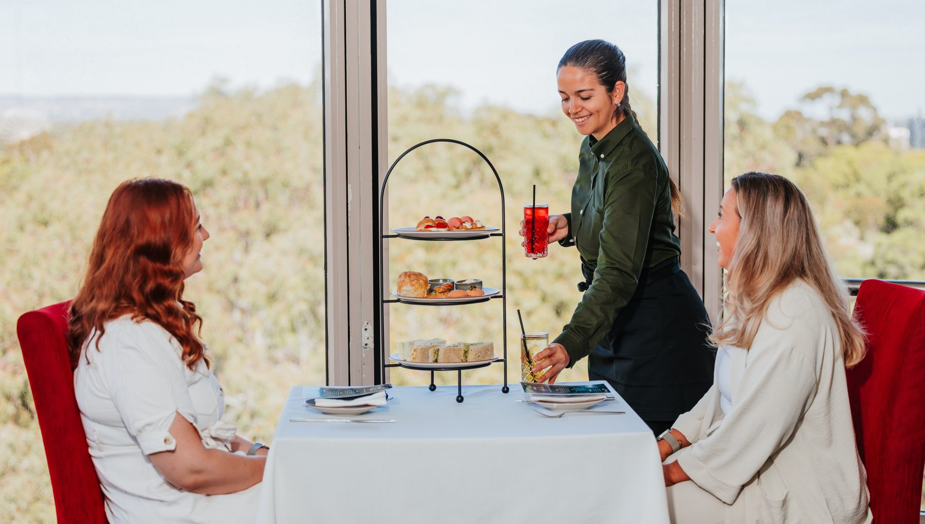 Two women sit at a table, smiling as a waitress serves drinks and a tiered platter of food, with greenery visible outside.
