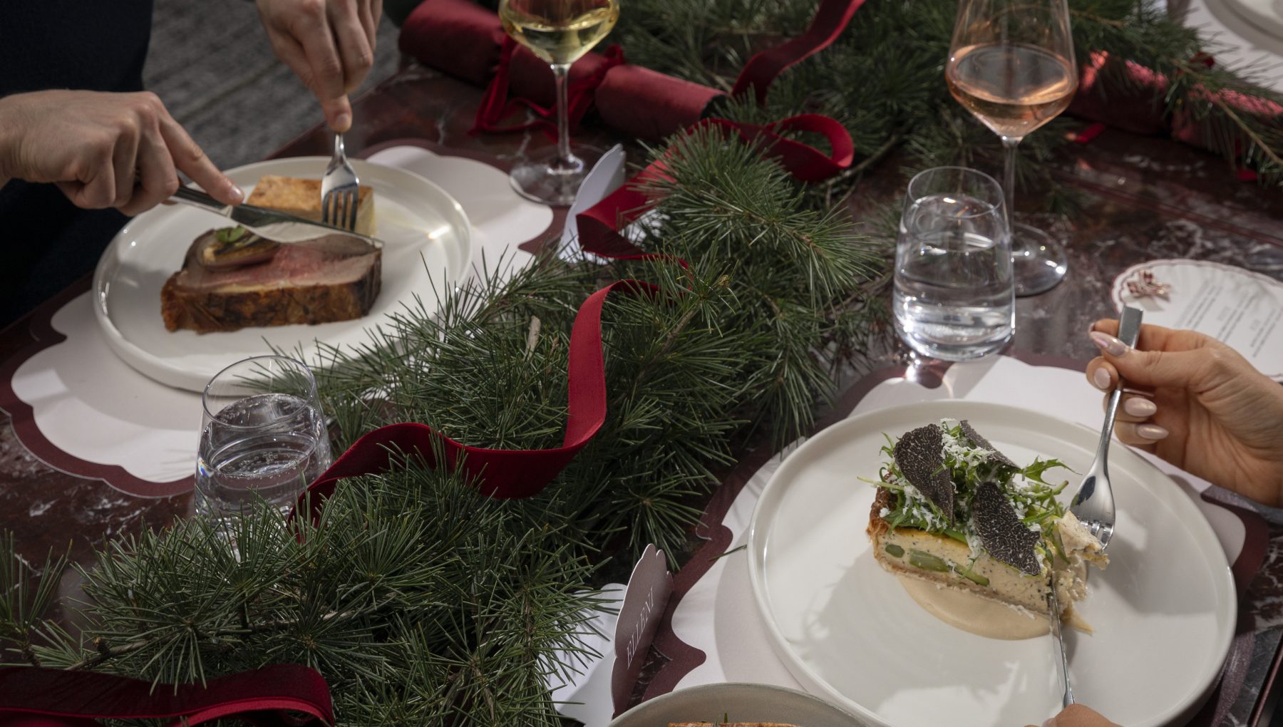 Hands serve a festive meal at a table decorated with evergreen branches and red ribbon, with plates, wine, and water glasses.