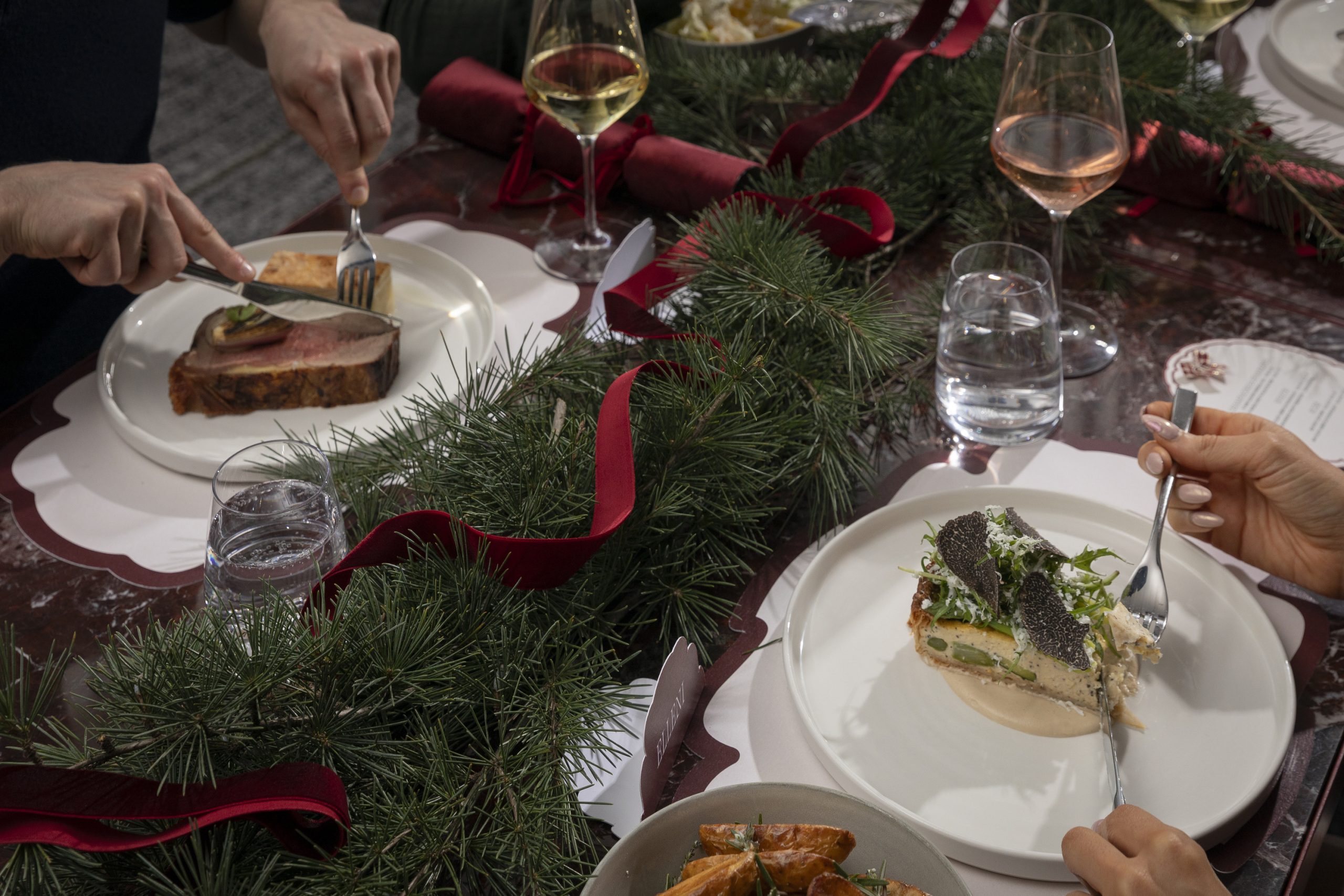 Hands serve a festive meal at a table decorated with evergreen branches and red ribbon, with plates, wine, and water glasses.
