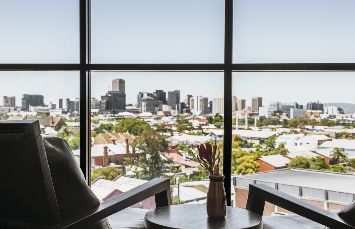 A view of a city skyline with modern buildings, framed by a large window, and a nearby chair with a small vase of flowers on a table.
