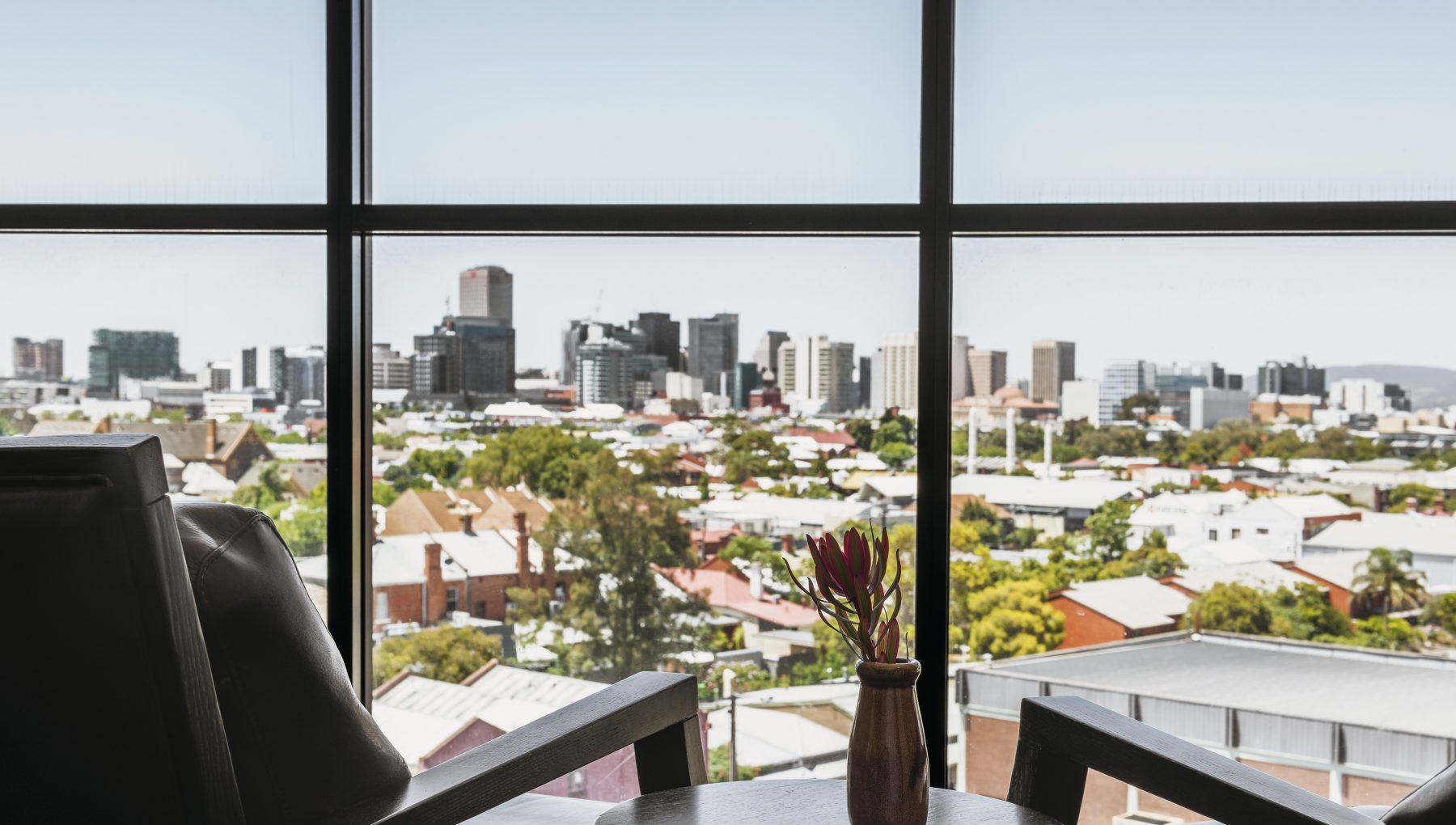 A view of a city skyline with modern buildings, framed by a large window, and a nearby chair with a small vase of flowers on a table.