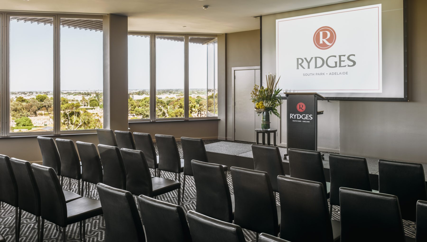 Conference room with black chairs facing a podium and screen, large windows showcasing a view of greenery outside.