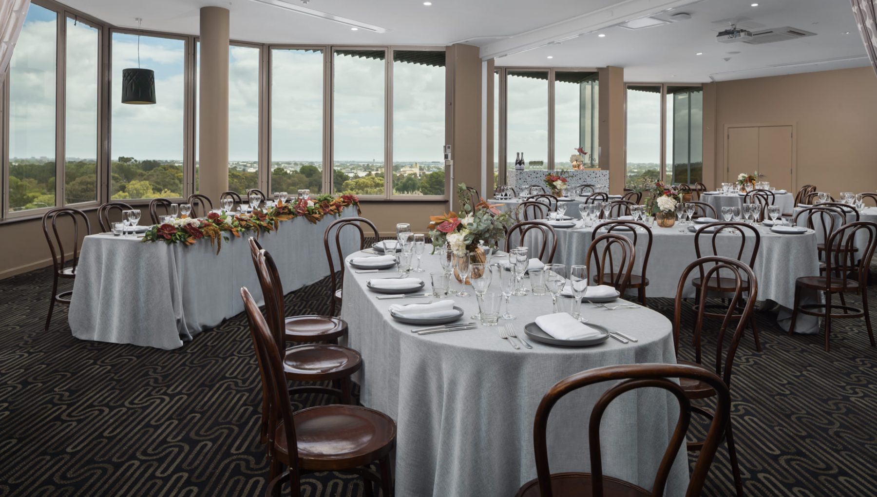 A banquet room with multiple round tables, each set with plates and glasses, features a floral arrangement. Large windows provide a view of greenery outside.