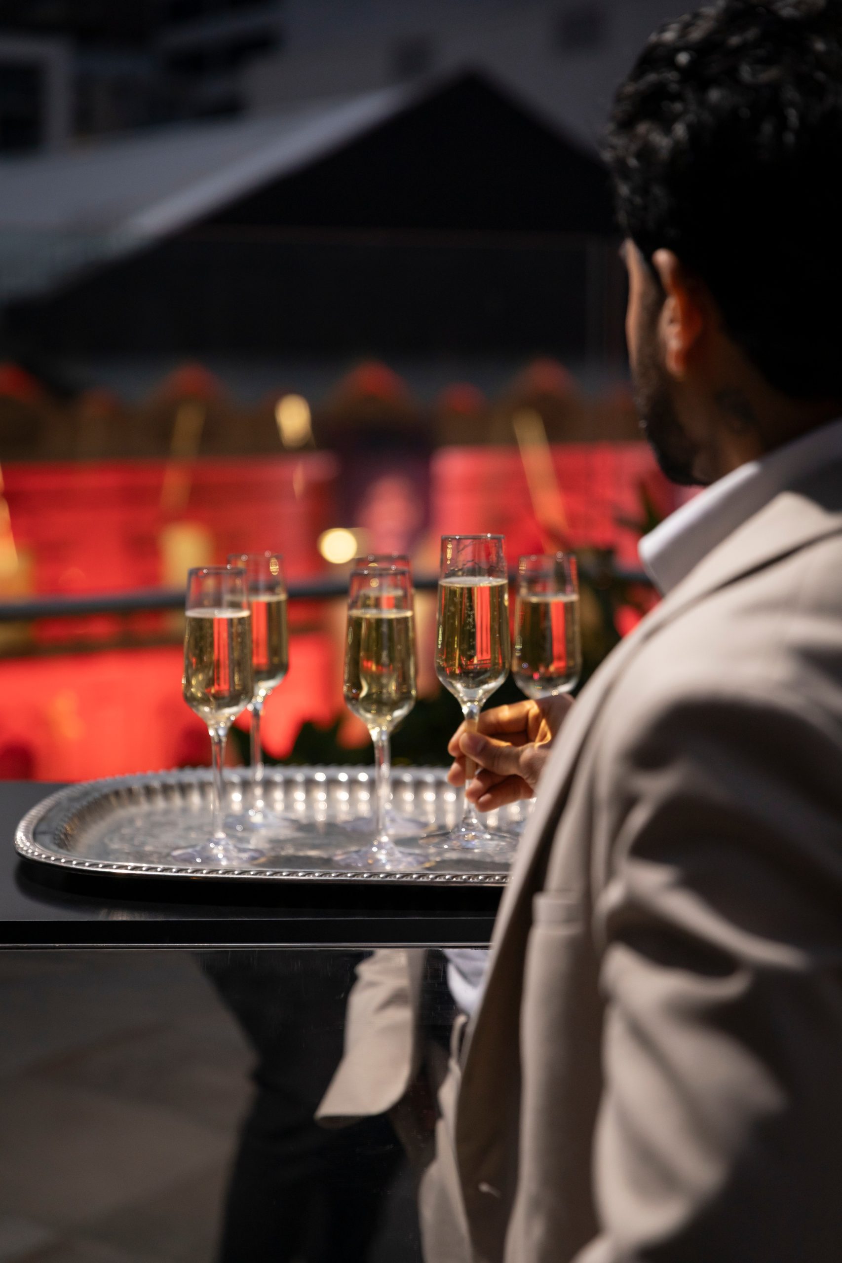 A person in a suit holds a tray with several glasses of champagne, overlooking a vibrant, illuminated background.