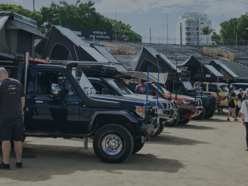 A row of off-road vehicles with rooftop tents is displayed at an outdoor event, with attendees interacting and exploring.