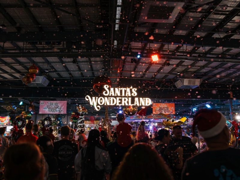 A festive indoor scene filled with people, decorated Christmas trees, and snow-like flakes, with a prominent sign reading "SANTA'S WONDERLAND."