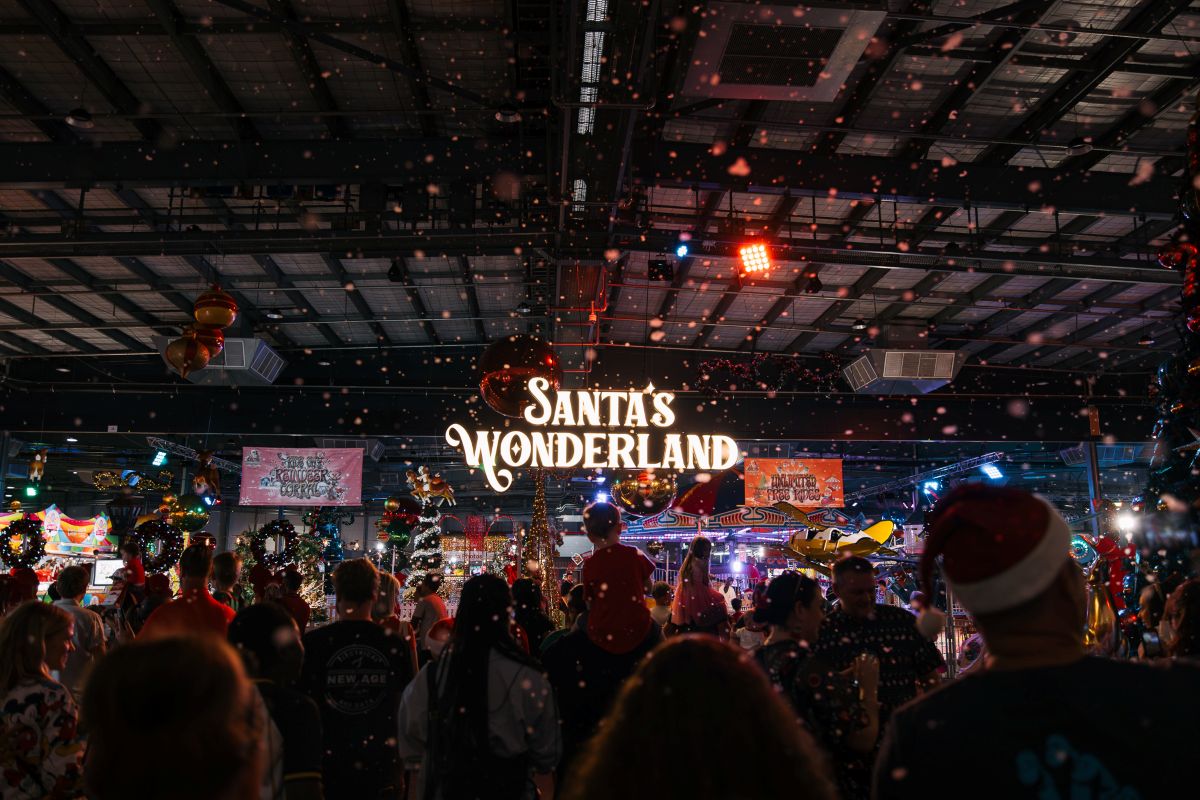 A festive indoor scene filled with people, decorated Christmas trees, and snow-like flakes, with a prominent sign reading "SANTA'S WONDERLAND."