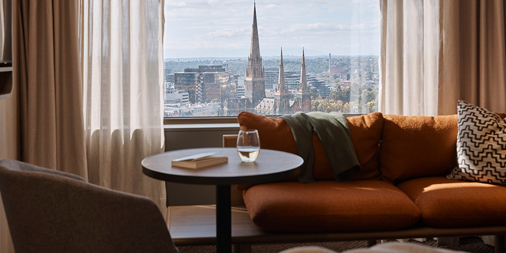 A cozy seating area with an orange couch and a round table, featuring a glass of water and a book, overlooking a cityscape with tall spires.