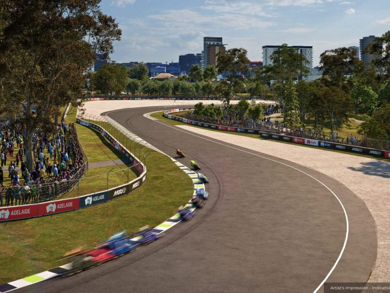 Motorcycles race around a tree-lined circuit curve, with crowds behind barriers and city buildings in the background.