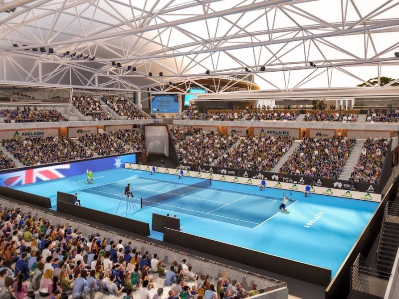 A crowded indoor tennis arena with players on a bright blue court beneath a white truss roof.