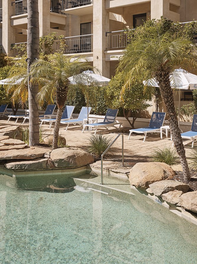 A clear pool with a sandy area, surrounded by palm trees and lounge chairs under umbrellas, beside a building.
