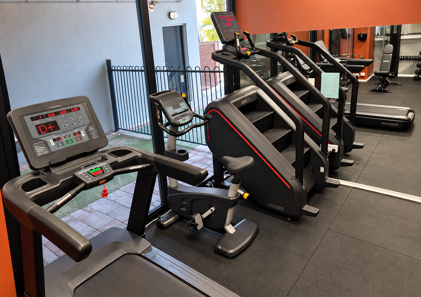 Cardio fitness equipment is displayed in a gym, including a treadmill, stair climbers, and a stationary bike. The environment has large windows and a modern design.