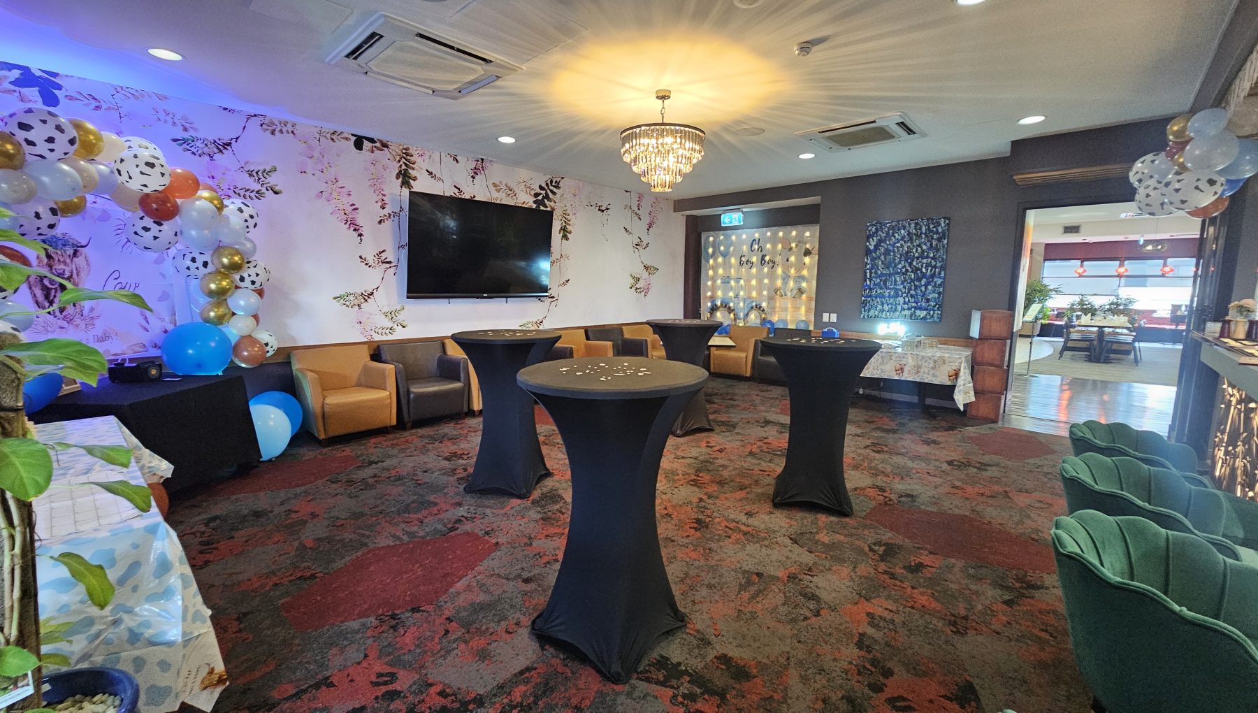 A decorated event room with balloon garlands, cocktail tables, lounge seating, floral walls, and a crystal chandelier.