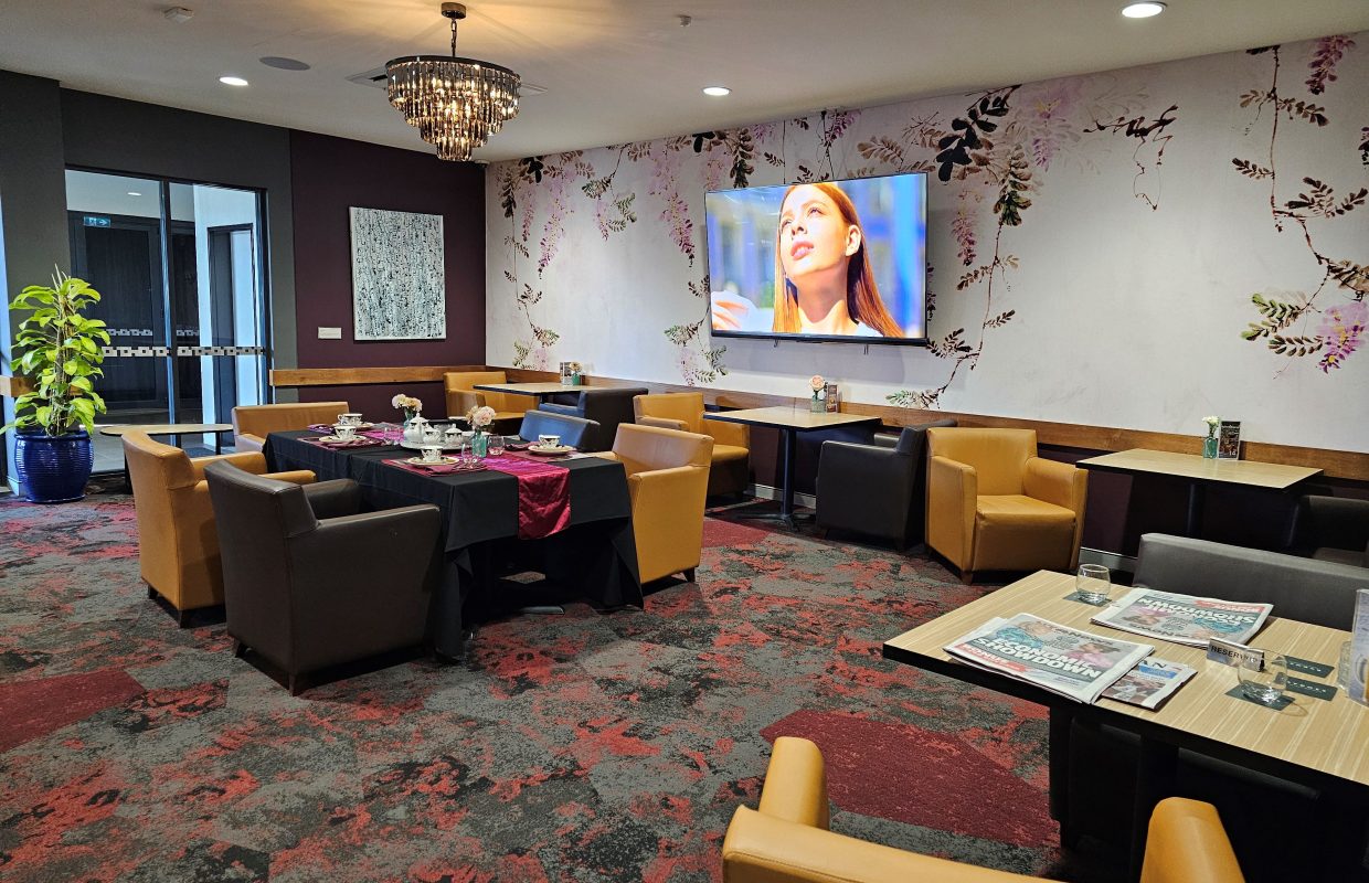 Restaurant lounge with tan and dark chairs, set tables, floral wallpaper, a chandelier, and a wall-mounted TV.