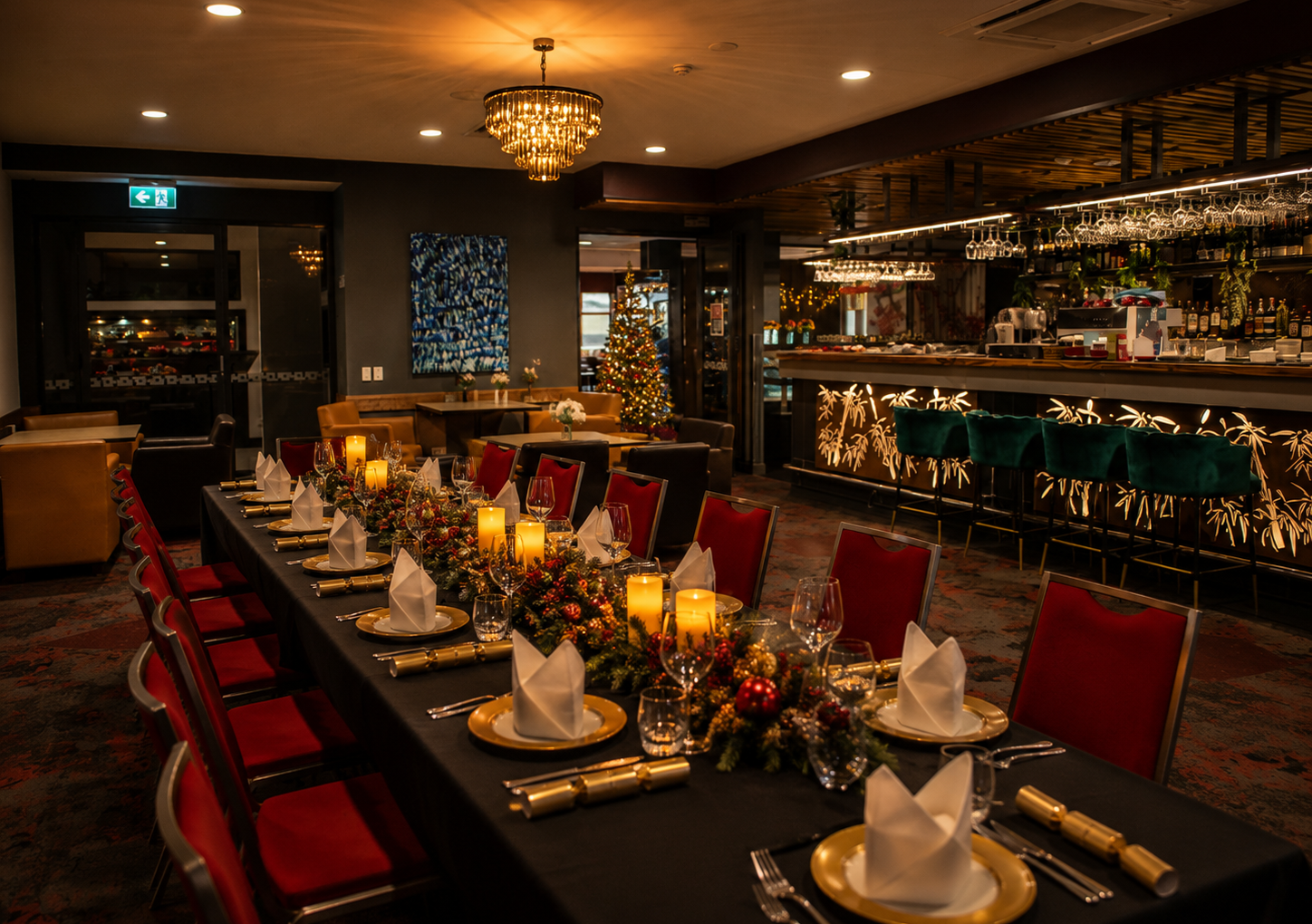 A warmly lit restaurant dining room with a long table set with candles, greenery, gold plates, and red chairs; a decorated Christmas tree stands near the bar.
