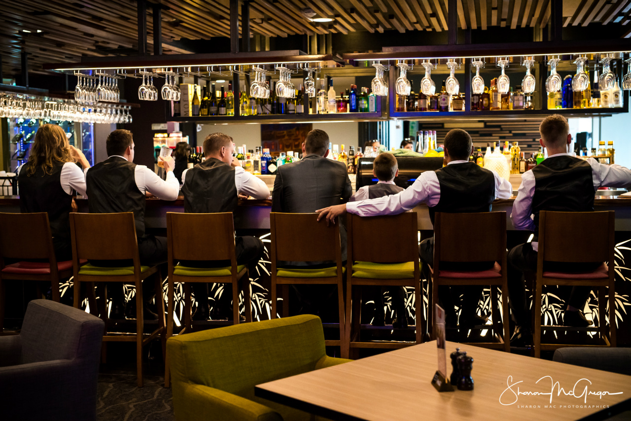 Patrons sit along a bar beneath hanging glasses, with liquor bottles lining the shelves behind them.