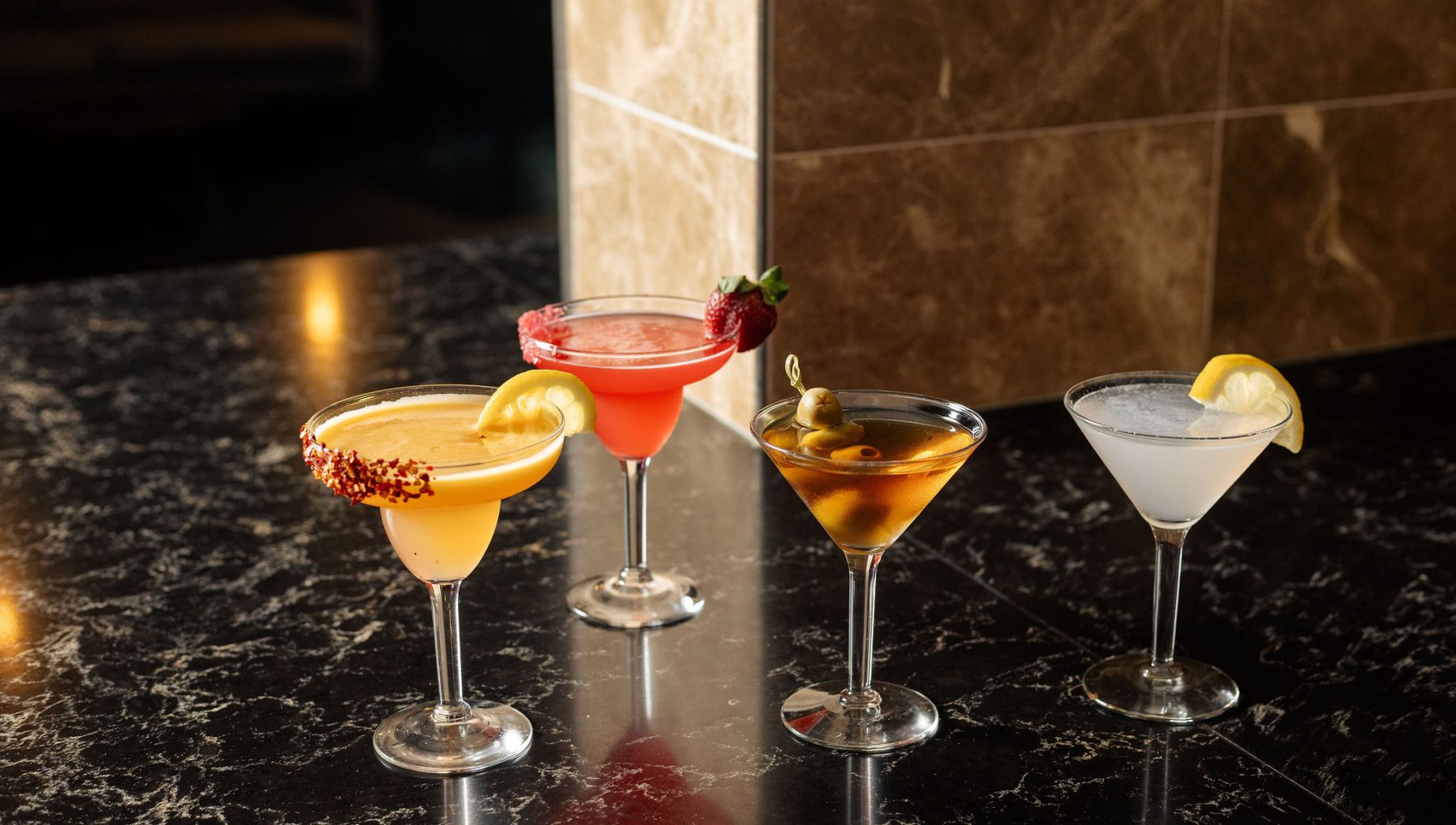 Four colorful cocktails are displayed on a dark marble surface, featuring garnishes like lemons and strawberries.