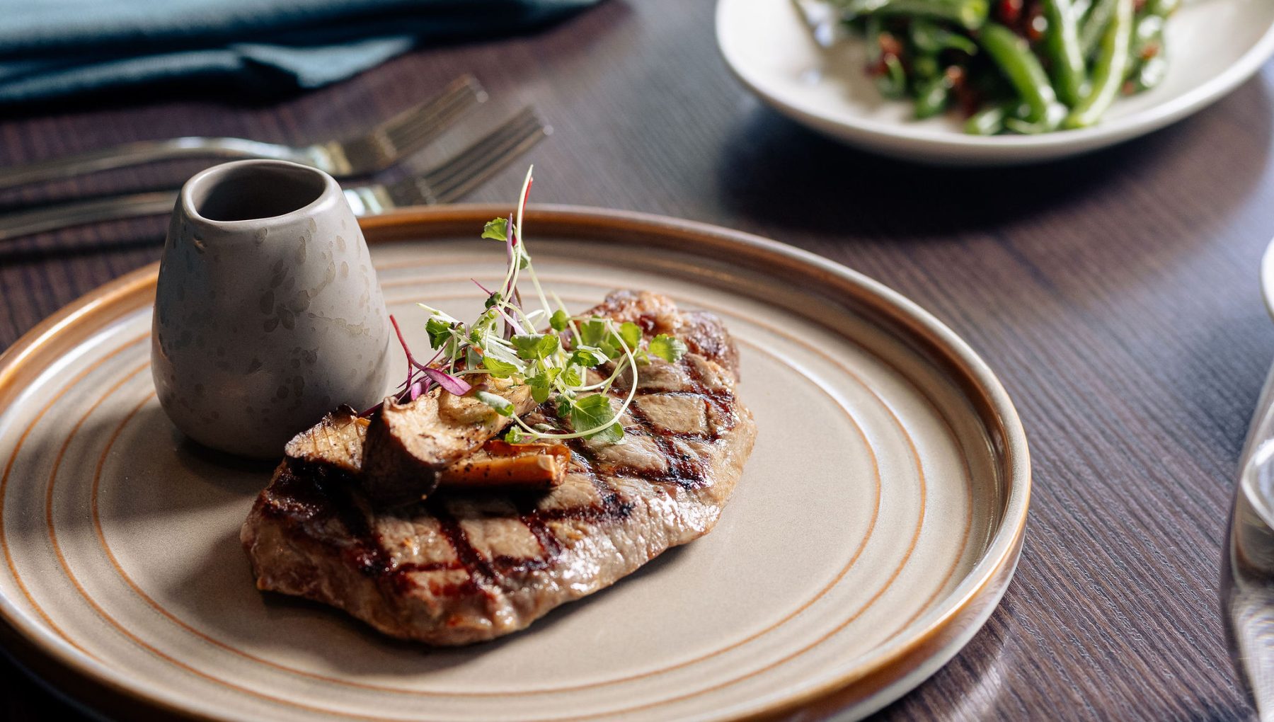 A grilled steak topped with microgreens and mushrooms is served on a beige plate, accompanied by a small sauce container.