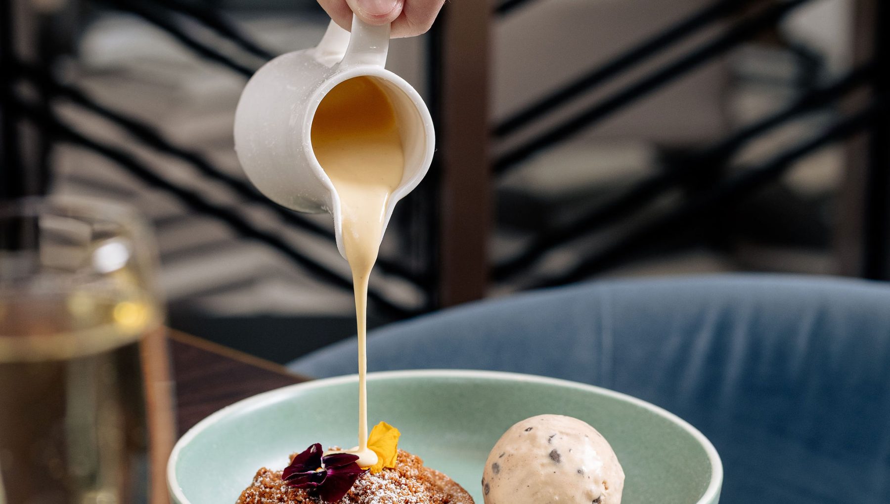 A hand pours sauce from a small white jug onto a dessert in a green bowl, featuring a scoop of ice cream and garnishes.