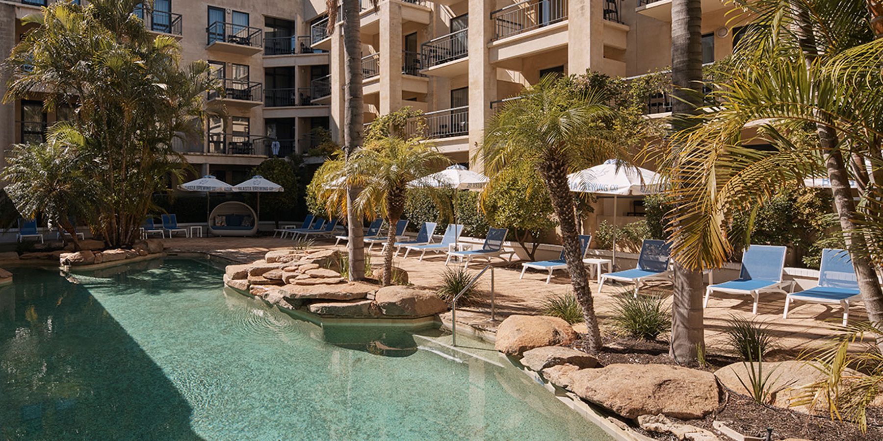 Apartment courtyard with a rock-edged swimming pool, palm trees, blue lounge chairs, and white umbrellas.