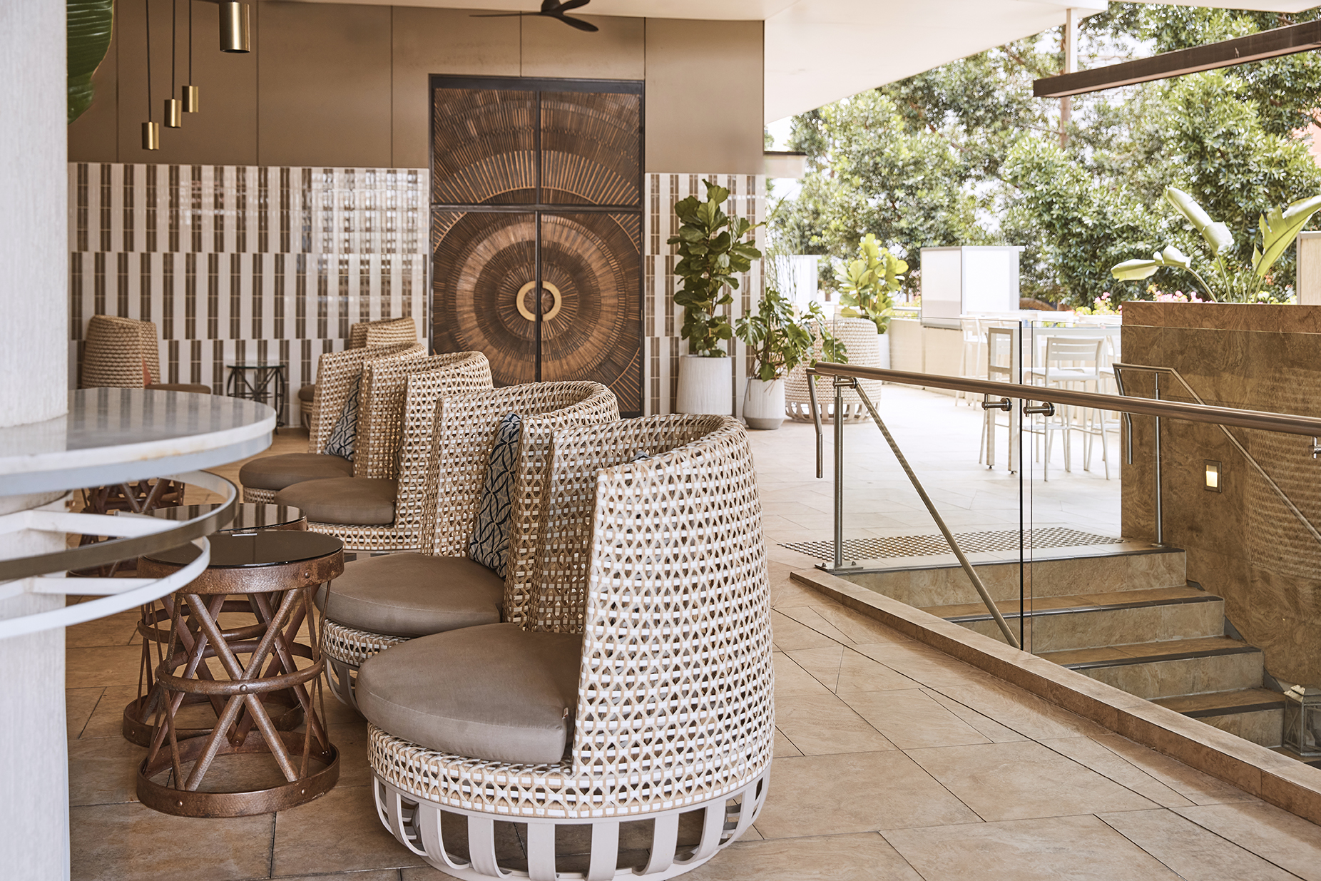 Stylish seating area with woven chairs and wooden tables, leading to an outdoor space with greenery and steps.