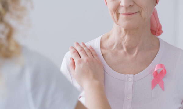 An older woman in a pink headscarf and pink ribbon rests her hand over another person’s hand on her shoulder.
